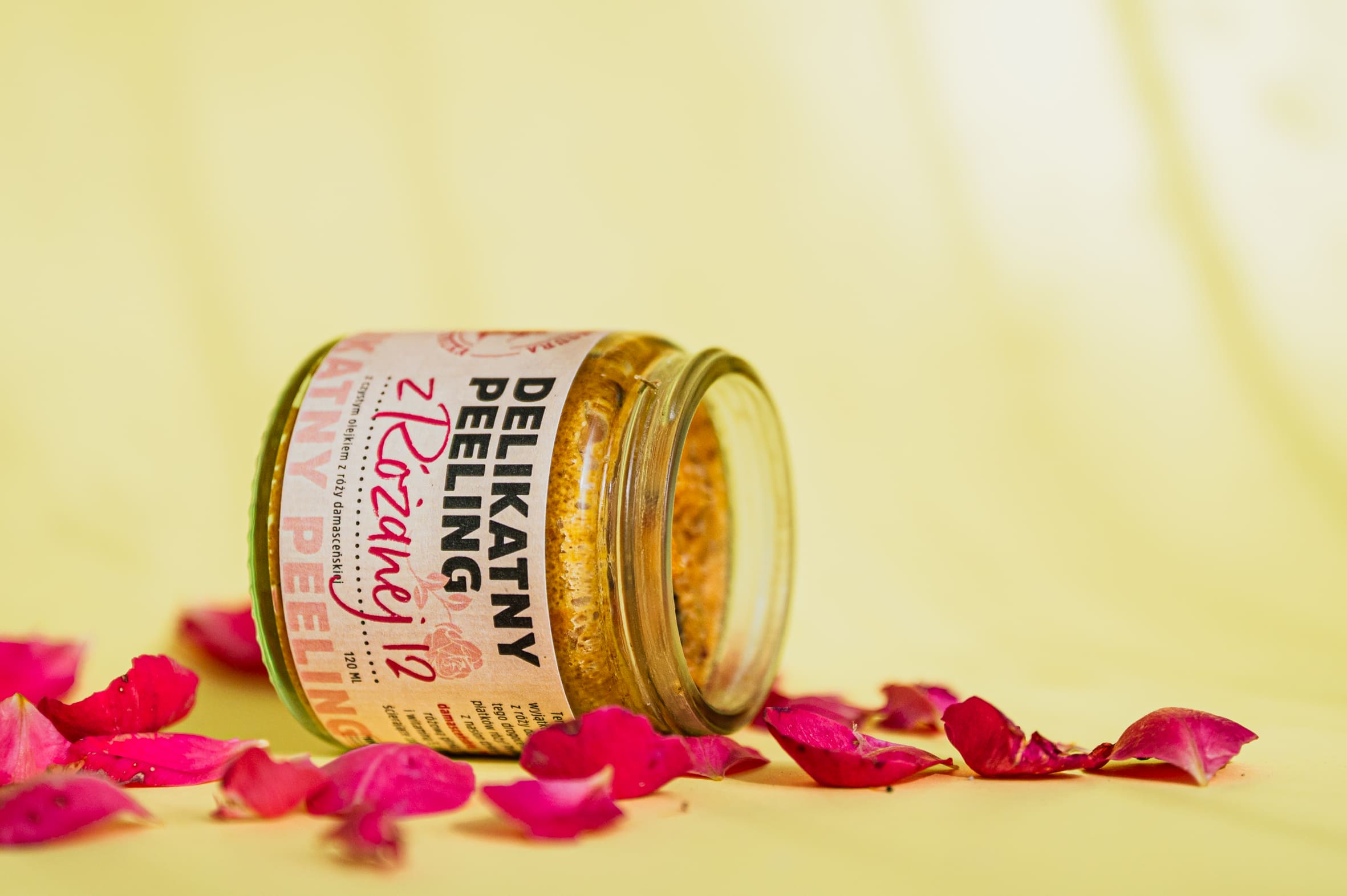 Słoik z peelingiem do ciała "Delikatny Peeling z Róży" leży na boku na żółtym tle, otoczony rozsypanymi, suchymi płatkami róży w różnych odcieniach różu. Peeling ma bursztynowy kolor.