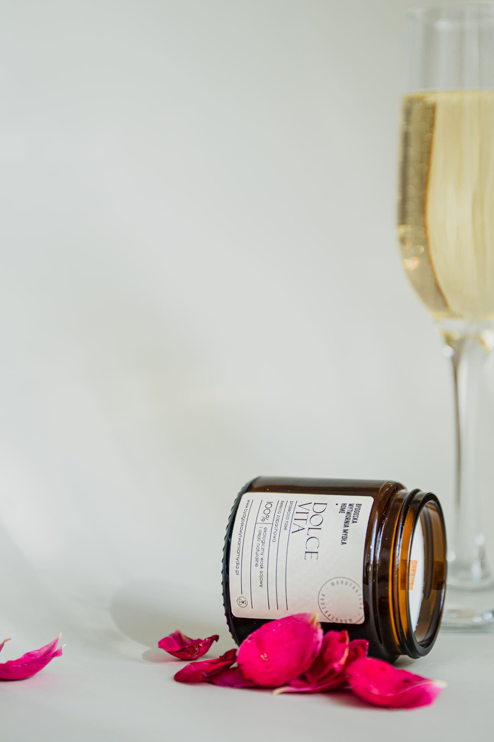 Pionowe ujęcie leżącej na boku, brązowej świecy sojowej "DATE NIGHT SPA" otoczonej różowymi płatkami róż, na białym tle. W tle widać górną część kieliszka wypełnionego szampanem lub innym złotym napojem.